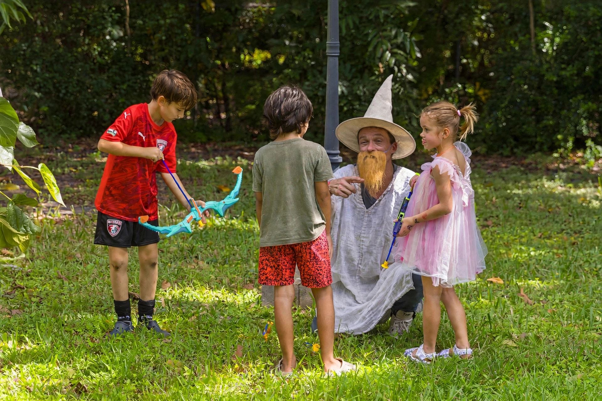 The bearded gnome in a pointed hat kneels beside young archers holding toy bows